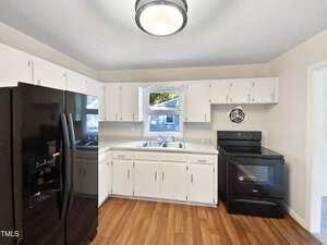 A kitchen with white cabinets, a black refrigerator, a black oven, wood-style flooring, a window above the sink, and a round ceiling light fixture.
