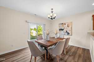 A dining room with a wooden table, six beige upholstered chairs, three white vases with decorative branches, a modern light fixture, abstract wall art, and a glass sliding door showing a view of greenery outside.