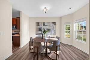 A dining area with a round wooden table, six gray upholstered chairs, a large abstract painting on the wall, and windows letting in natural light. The space has light walls and wood-style flooring.