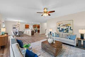 Spacious open-concept living and dining area with wood flooring, neutral walls, and a ceiling fan. The room features modern furniture, decorative plants, artwork, and a view into the adjacent kitchen.