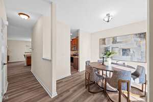 A dining area with a round wooden table, six gray upholstered chairs, a potted plant centerpiece, and a large abstract painting on the wall. The space has light beige walls and wood-style flooring.