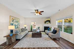 A living room with a beige sofa, two armchairs, a wooden coffee table, abstract wall art, a ceiling fan with lights, a fireplace, large rug, and windows letting in natural light. The floor is wood-patterned.