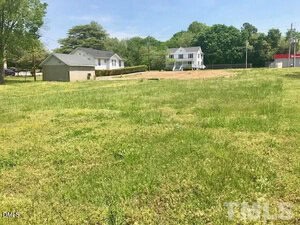 A grassy, empty lot with a few small houses and trees in the background under a clear sky. A gray shed is visible to the left, and there are power lines and a red-roofed building on the far right.