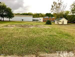 A grassy lot in Oxford at 209 Broad Street features a concrete slab at its center, bordered by trees. In the background are a white warehouse-like building, a small light-colored shed, and a brick building with blue doors and windows.