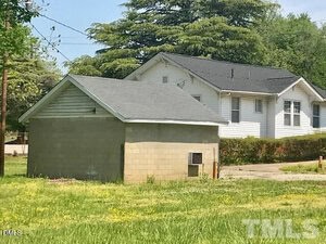 A small cinder block structure with a gray roof stands on grass near a white house with black shingles. Trees and a driveway are in the background. The image has a TMLS watermark in the corner.