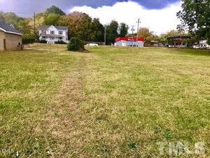 A grassy vacant lot with a faint path leading toward a white house on the left and a red-roofed building in the background. Trees with early autumn foliage line the edges under a cloudy sky.