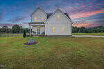 A two-story white house at 90 Eagle Chase Drive, Youngsville, features a covered front porch with rocking chairs, set on a large, neatly mowed lawn at sunset. A stone surrounded by mulch graces the front yard with trees in the background.