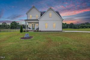 A two-story white house at 90 Eagle Chase Drive, Youngsville, features a covered front porch with rocking chairs, set on a large, neatly mowed lawn at sunset. A stone surrounded by mulch graces the front yard with trees in the background.
