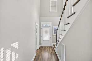 A hallway with light-colored walls, wooden flooring, a white front door with glass panels, and a staircase with white risers and dark wooden handrails leading upward on the right side.