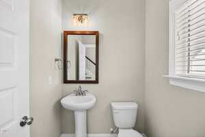 A small bathroom with a pedestal sink, a wall-mounted mirror with a wooden frame, a toilet, a towel ring, and a window with white blinds. The walls are painted a light beige color.