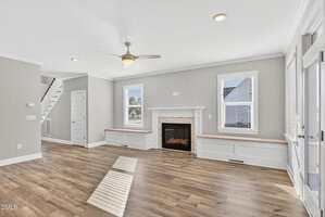 A bright living room with light gray walls, wood flooring, a ceiling fan, a white fireplace between two windows, built-in bench seating, and a staircase leading upstairs.