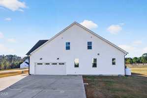 A large white building with a gable roof, three garage doors, and several windows sits on a concrete driveway. There is a propane tank on the right side and grassy areas surrounding the driveway. Trees are visible in the background.