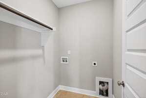 A small laundry room with beige walls, a tile floor, a wooden shelf with a hanging rod, electrical and water hook-ups for a washer and dryer, and a white door partially visible on the right.