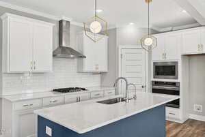 Modern kitchen with white cabinets, stainless steel appliances, a central island with a sink, two geometric pendant lights, subway tile backsplash, and light wood flooring.