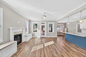 Bright open-concept living area with wood flooring, a white fireplace, built-in benches, and large windows. The kitchen features a blue island, pendant lights, and white cabinetry. French doors lead outside.