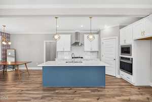 Modern kitchen with a blue island, white cabinets, stainless steel appliances, and wood flooring. Pendant lights hang above the island, and a dining area with a table and chairs is visible to the left.