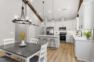 A modern kitchen and dining area with white cabinets, stainless steel appliances, a center island with three wooden stools, light wood flooring, pendant lighting, and a dining table with four chairs and a flower vase.
