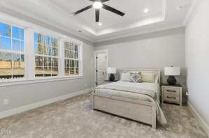 A neatly made bed with two nightstands and lamps sits in a carpeted bedroom with light gray walls. Large windows on the left let in natural light, and a ceiling fan is centered on the tray ceiling.