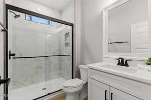 Modern bathroom with white cabinets, a white countertop, black fixtures, a toilet, and a glass-enclosed shower with marble-patterned tile. A small plant and rolled towel sit on the counter.