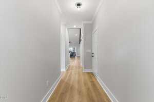 A hallway with light-colored walls, wood flooring, white trim, and ceiling light leads to an open living area. A closed white door is on the right side of the hallway.