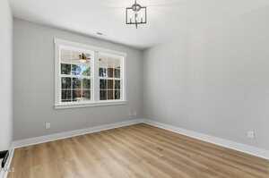 A bright, empty room with light wood flooring, gray walls, white trim, a ceiling light fixture, and a double window showing trees and a ceiling fan outside.