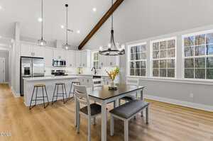 A modern kitchen and dining area with white cabinets, stainless steel appliances, and a dining table with chairs and a bench. Large windows let in natural light, and pendant lights hang from the ceiling.