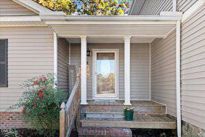 A covered front porch with beige siding, white trim, and a wooden ramp leading to a glass-paneled door. A potted plant and a "WELCOME" sign sit near the entrance. Shrubs are on the left side.