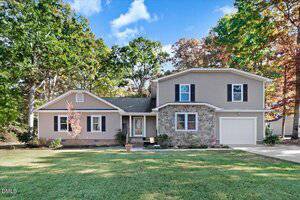 A two-story house with beige siding and stone accents at 5014 Country Day Drive, Zebulon. Features an attached single-car garage, gabled roof, landscaped front yard with green grass and potted plants, surrounded by tall trees under a partly cloudy sky.