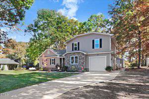 Two-story suburban house with beige siding and stone accents, attached single-car garage, concrete driveway, and a well-maintained front yard with shrubs and trees. The sky is clear with some clouds.