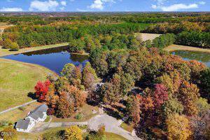 Aerial view of a rural property with a house, a dirt driveway, surrounding trees in fall colors, a large pond, open fields, and dense forest in the background under a blue sky with scattered clouds.