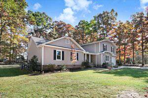 A tan, two-story suburban house with white trim, surrounded by trees with autumn foliage. The house has a front porch, a gabled roof, and a well-maintained lawn under a blue sky with scattered clouds.