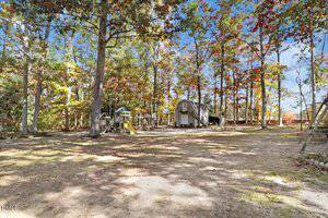 A large, partially shaded backyard with scattered trees, a wooden playset with slides on the left, and a white barn-style shed with a curved roof in the center. The ground is mostly dirt with patches of grass.