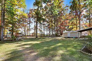 A spacious backyard with green grass, surrounded by tall trees with autumn foliage. There is a wooden playset on the left, a small garden bed, and a white shed and covered patio area on the right side.