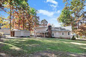 Two-story house with brick chimney, surrounded by trees with autumn foliage. A detached garage or shed sits to the left. The backyard is grassy and partially shaded, under a blue sky with scattered clouds.
