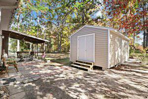 A light-colored shed with double doors sits on a sandy area next to a covered outdoor patio with a dining table and chairs. The yard is surrounded by tall trees with green and autumn-colored leaves.