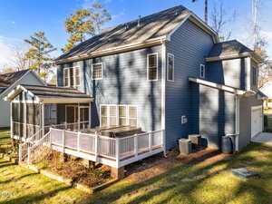 A two-story blue house with white trim features a screened porch, large raised deck with a hot tub, two air conditioning units, a rain barrel, and a two-car garage. The yard is grassy with some trees and landscaping.