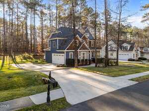 A two-story house with blue siding and brown accents sits next to a similar neighboring home. Both have attached garages and well-maintained lawns. Tall trees line the background and a black mailbox is by the curb.