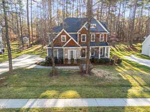 A two-story house with a front porch and gabled roof is surrounded by trees and a lawn. A driveway curves on both sides of the house, and a sidewalk runs along the street in front.
