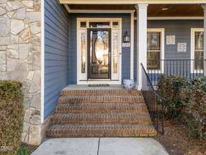 A brick staircase with six steps leads to a glass front door of a blue house with white trim. A black railing, potted plant, and house number 105 are visible. Shrubs and a stone wall frame the entry.