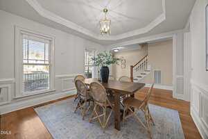 A dining room with a wooden table, six wicker chairs, a blue and white rug, a black vase with green branches, hardwood floors, white walls, large windows, and a gold chandelier. Stairs are visible in the background.