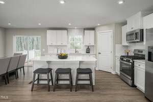 A modern kitchen with white cabinets, stainless steel appliances, a center island with three stools, wood flooring, and a dining table partially visible to the left. Large windows let in natural light.