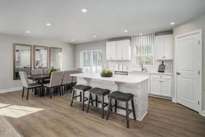 Modern kitchen with white cabinets and countertops, a central island with three stools, and a dining area with a table and upholstered chairs. Large windows provide natural light, and wood floors run throughout the space.