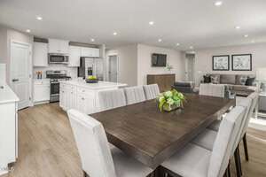 Open-concept kitchen, dining, and living area with white cabinets, stainless steel appliances, and wood-style flooring. A dark wood dining table with upholstered chairs sits near a living room with a gray sofa and framed art.