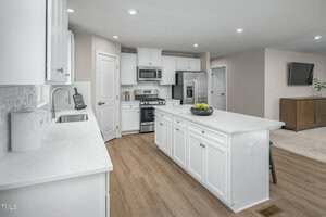 Modern kitchen with white cabinets, stainless steel appliances, a gas stove, a large white island with a bowl of green apples, wood flooring, and an open view into a living area with a TV.