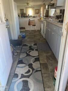 A view of a narrow kitchen with gray cabinets, countertop appliances, and a patterned rug on a tiled floor. Boxes, water bottles, and various items are scattered throughout the space. Sunlight enters from windows.