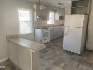 A kitchen with gray cabinets, white appliances including a stove and refrigerator, a light fixture, a countertop peninsula, tiled floor, and two windows letting in natural light.