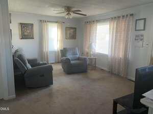 A living room with light-colored walls, beige carpet, two gray recliner chairs, a side table with a lamp, framed artwork, two windows with curtains, and a ceiling fan with lights. A TV is visible in the foreground.