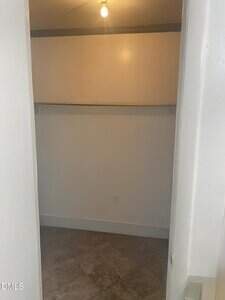 A small, empty closet with white walls, a single exposed light bulb on the ceiling, a wooden shelf with a hanging rod, and an electrical outlet on the back wall. The floor appears to be tile or linoleum.