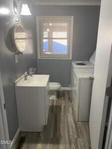 A small bathroom with gray walls and wood-look flooring includes a white vanity with a sink and mirror, a toilet, and a top-loading washing machine next to a window with blinds.