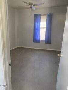 An empty room with gray carpet, light gray walls, a ceiling fan, and a window covered by blue curtains letting in sunlight. The door is partially open, showing the simple interior.